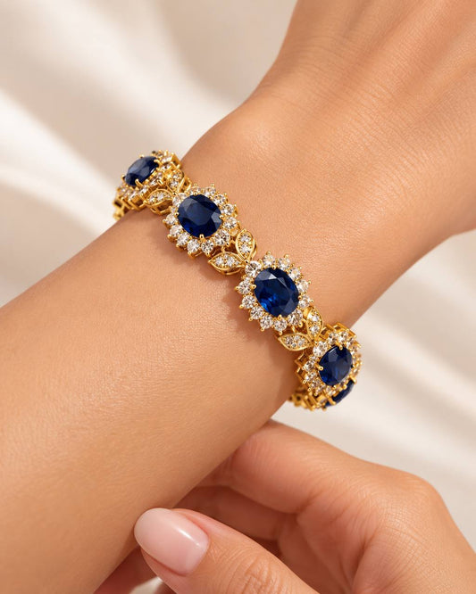 A close-up of a gold bracelet with oval blue gemstones and small clear stones, worn on a person's wrist against a soft light background.