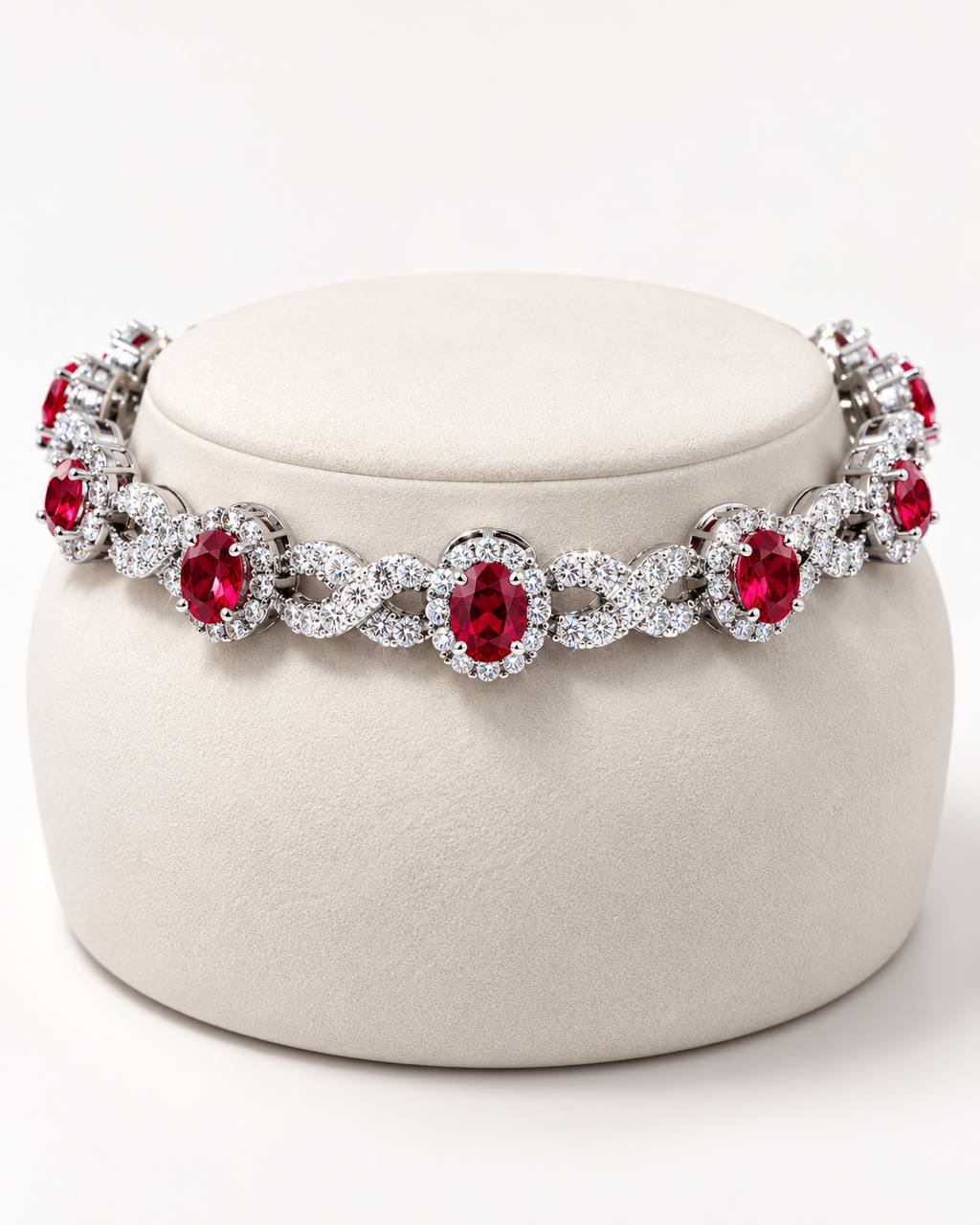 A silver-toned tennis-style bracelet with oval red gemstones and surrounding clear stones, displayed on a round cream jewelry stand against a light gray studio background.