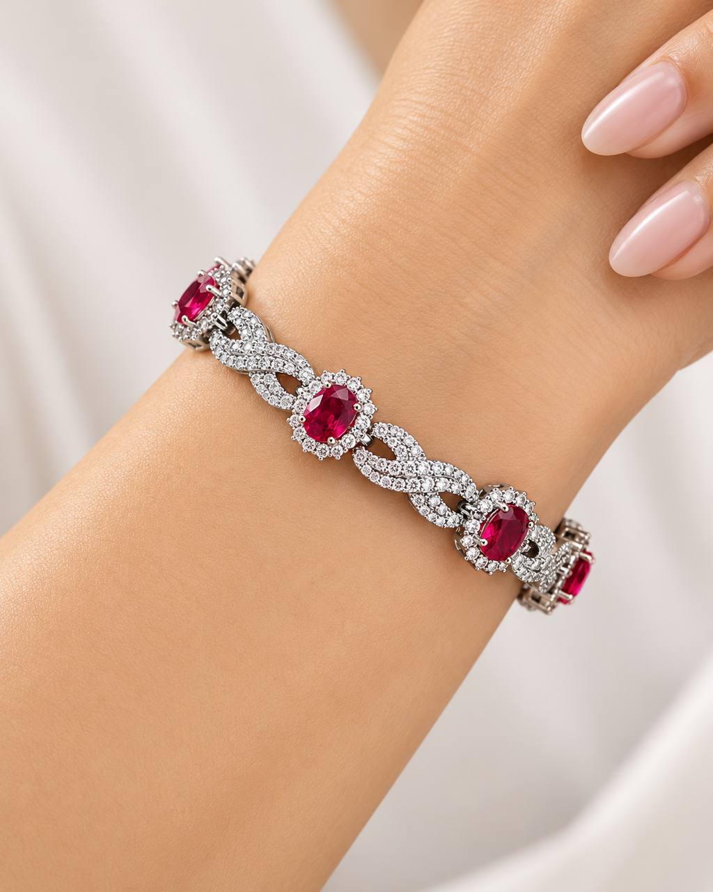 A close-up of a silver-toned bracelet with oval red gemstones and surrounding clear stones, worn on a person's wrist against a soft white background.